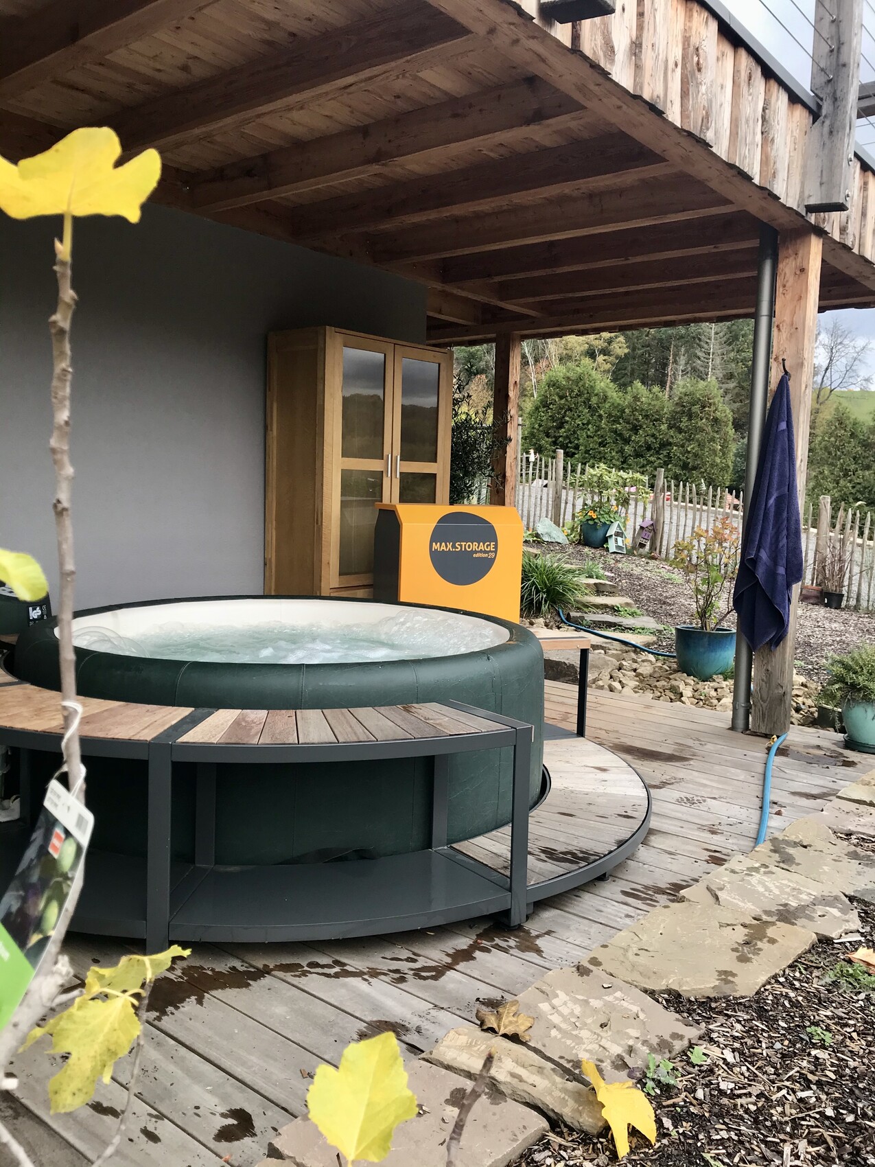 Rounded outdoor hot tub on wooden deck under pergola; yellow MAX STORAGE box in background.