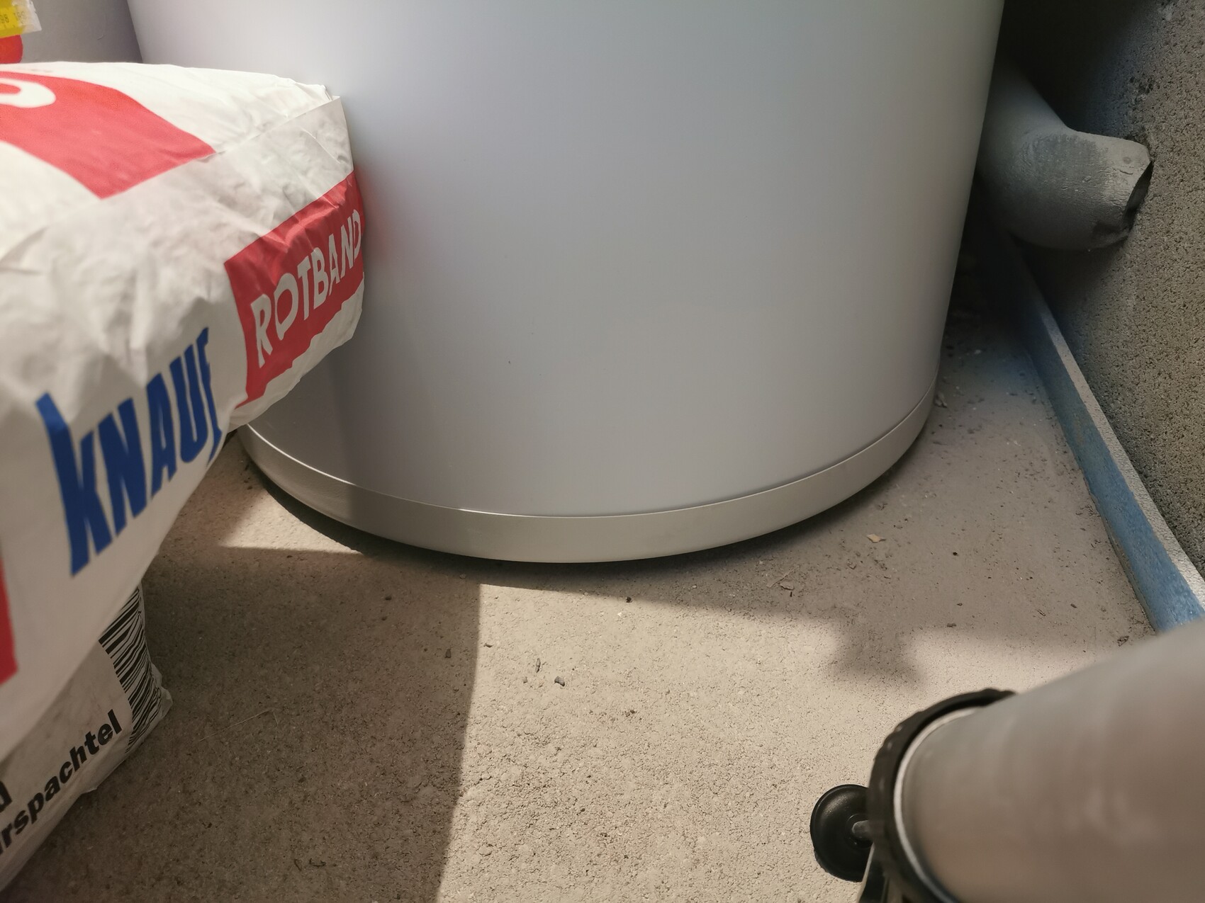 Large white cylinder boiler standing on dusty floor next to a Redband cement bag in the basement.
