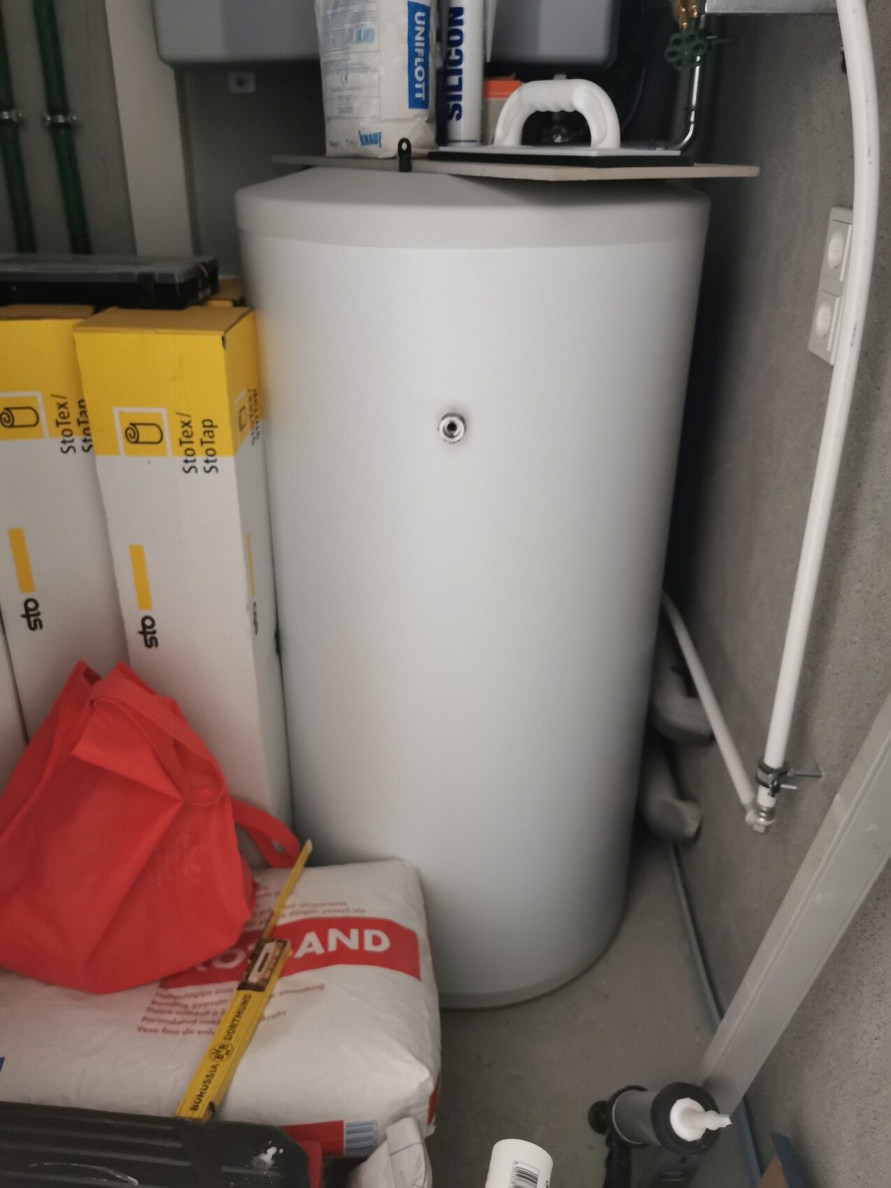 White hot water boiler in a storage room, surrounded by yellow cardboard boxes and a red bag.