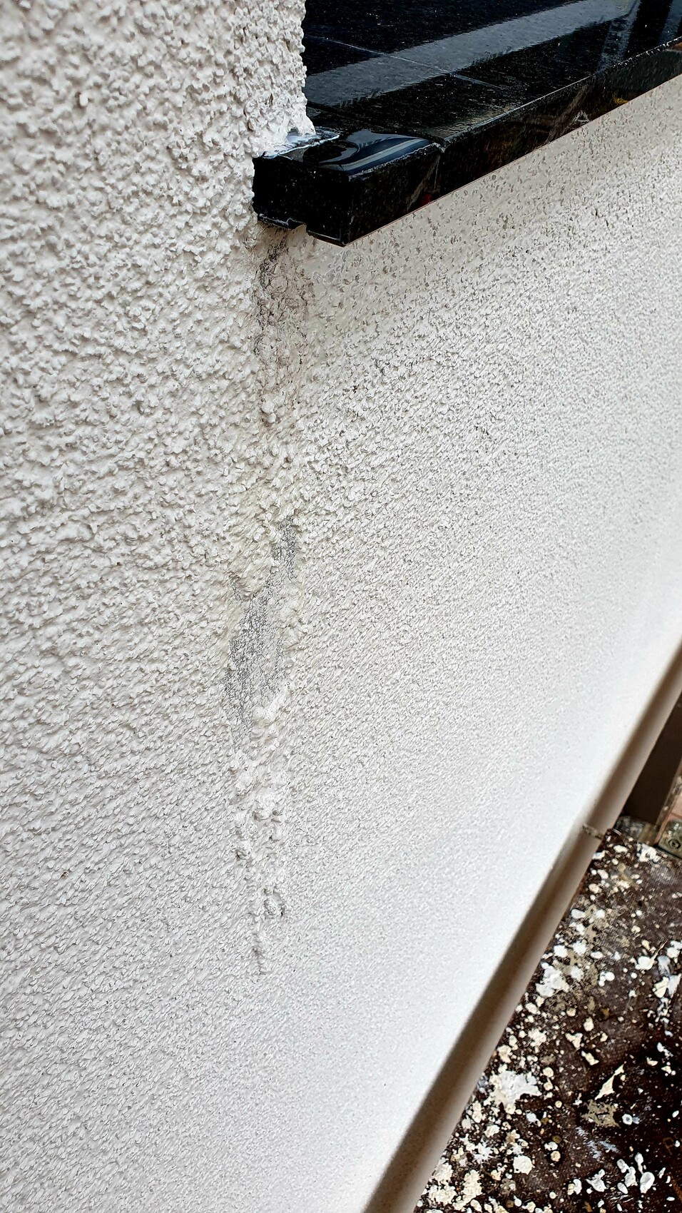 Exterior wall with rough render texture, black stone strip above, moisture marks and dust on the ground.