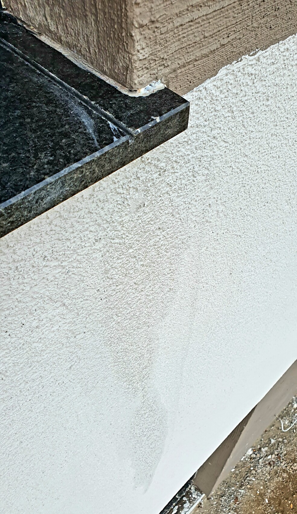 Exterior wall corner: black granite threshold over white render wall, sealing compound visible.