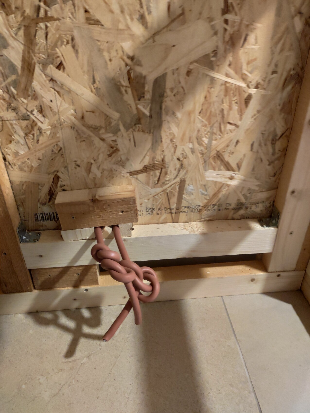 OSB wall with timber frame; below a wooden block with two brown cables, loosely knotted.