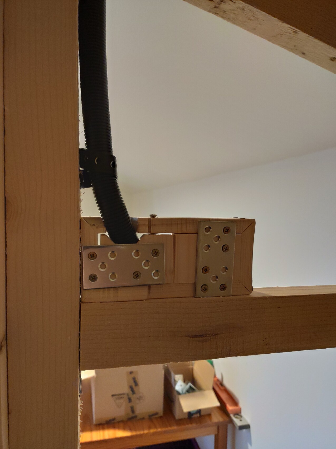 Timber frame with metal angle brackets and black cable conduit; background cardboard boxes.