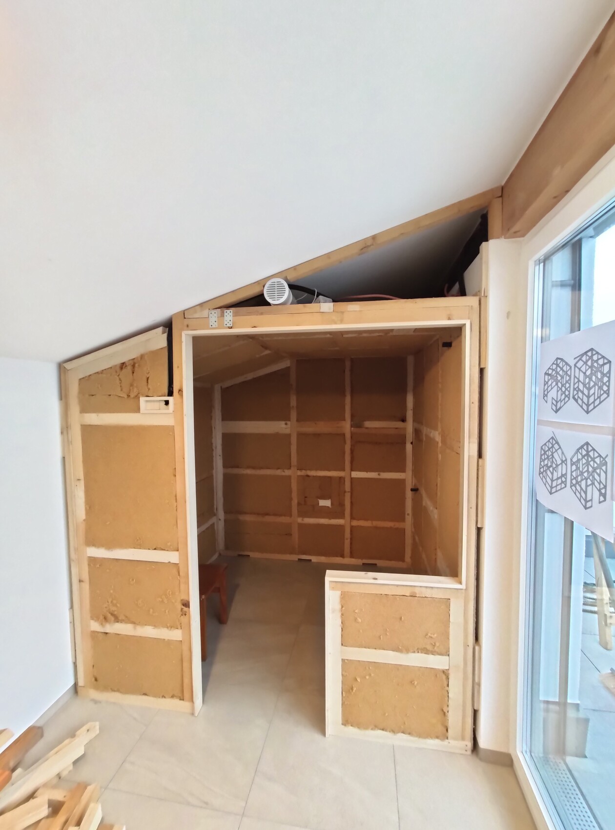 Unfinished interior under sloping roof, timber frame, open cavities, small bench at front.
