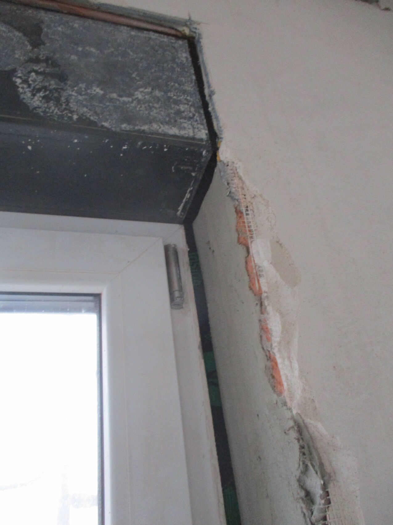 Window frame next to damaged wall; exposed brick, plaster residues, and wire mesh visible.