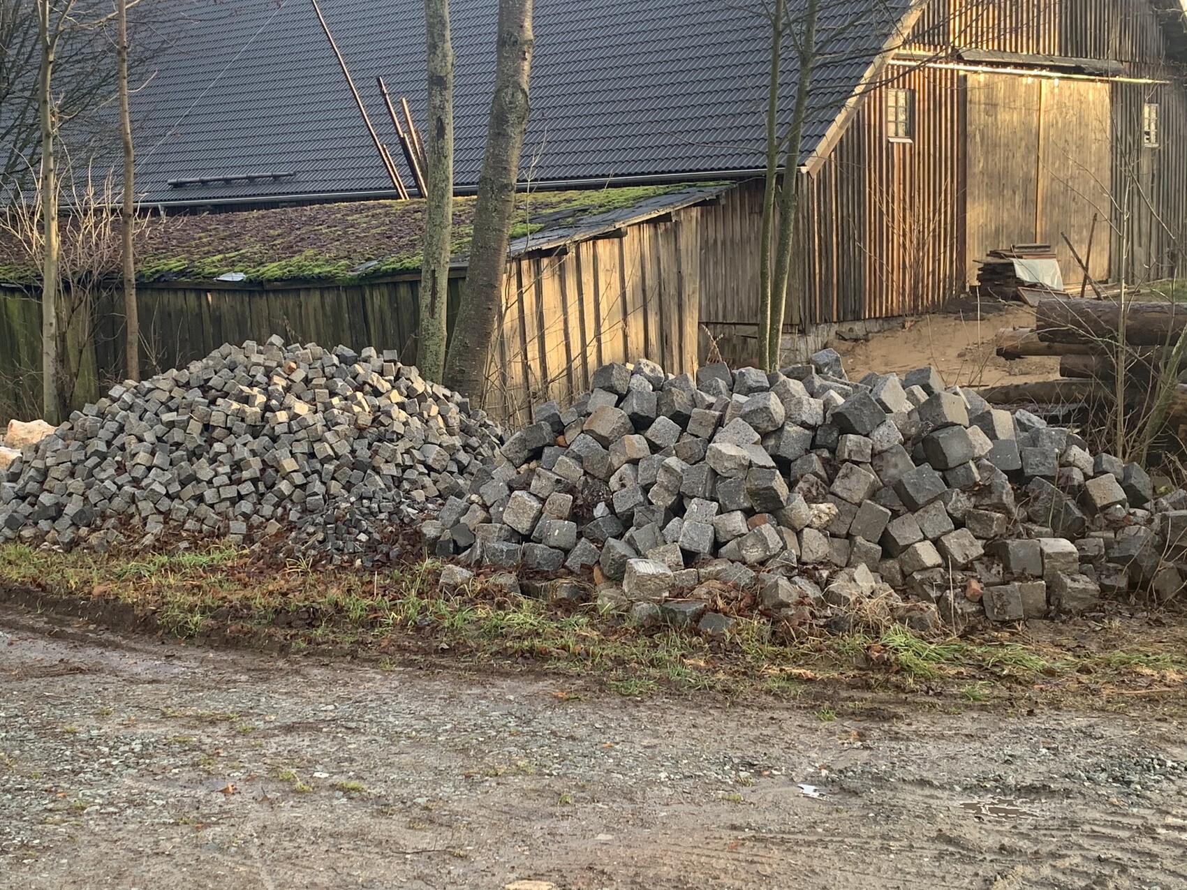 Two piles of gray cobblestones in front of a wooden cabin; trees stand in the background.
