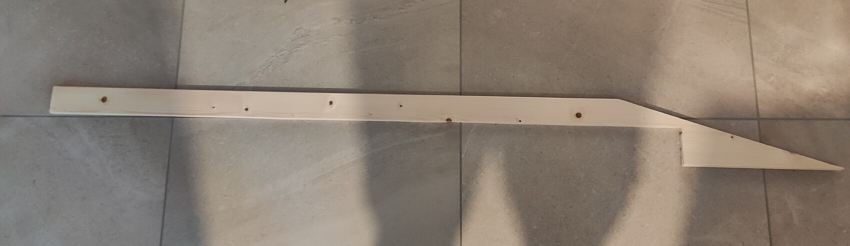 Long wooden batten with a wedge-shaped tip on the right end lying on tiled floor.