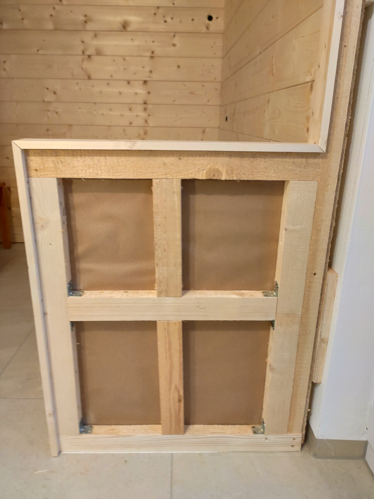 Wooden frame with vertical central support and cross braces in front of wood wall; metal brackets at the bottom.