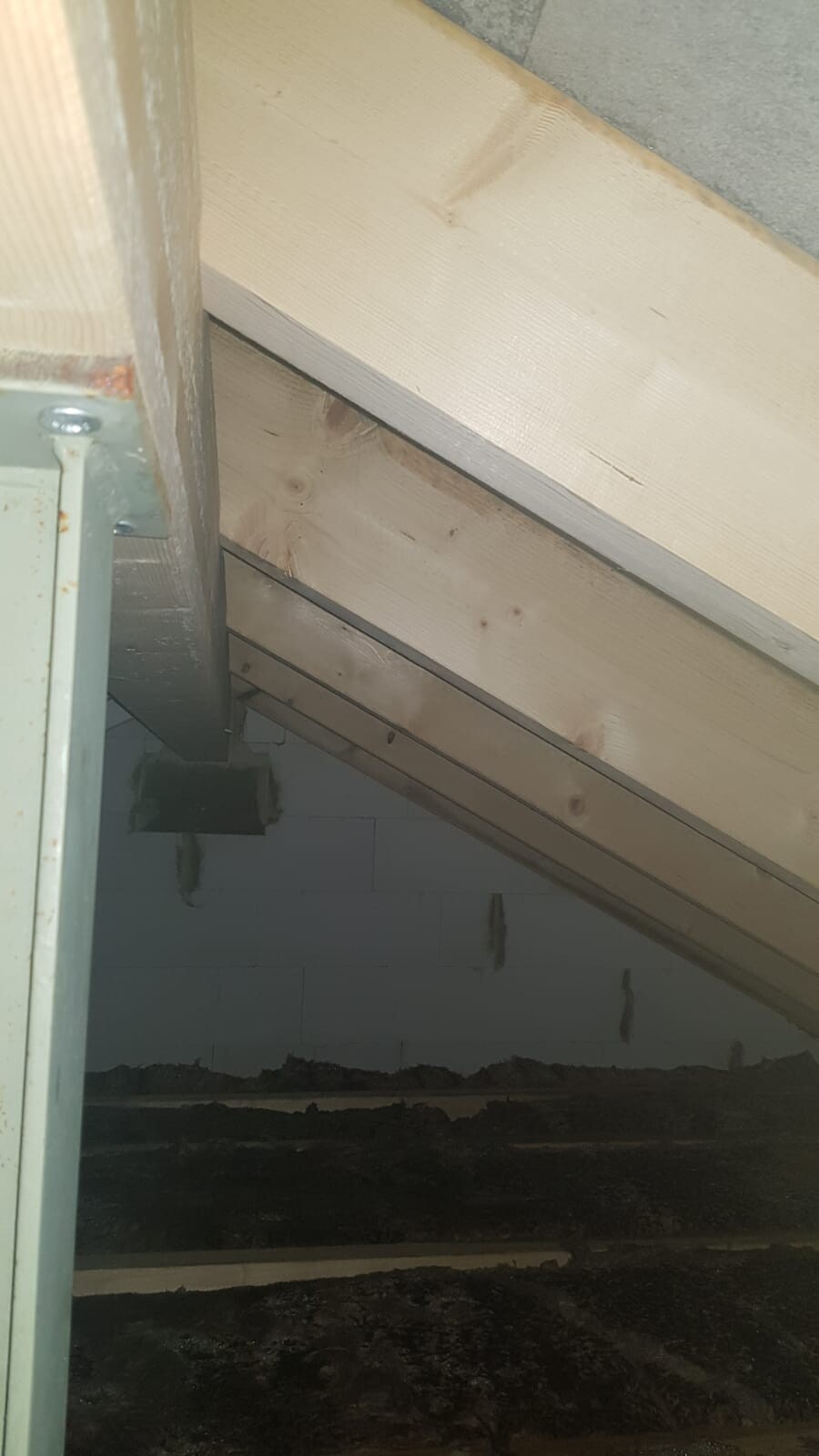 Sloping roof frame made of light wooden beams, metal bracket on the left, concrete wall, dark floor.