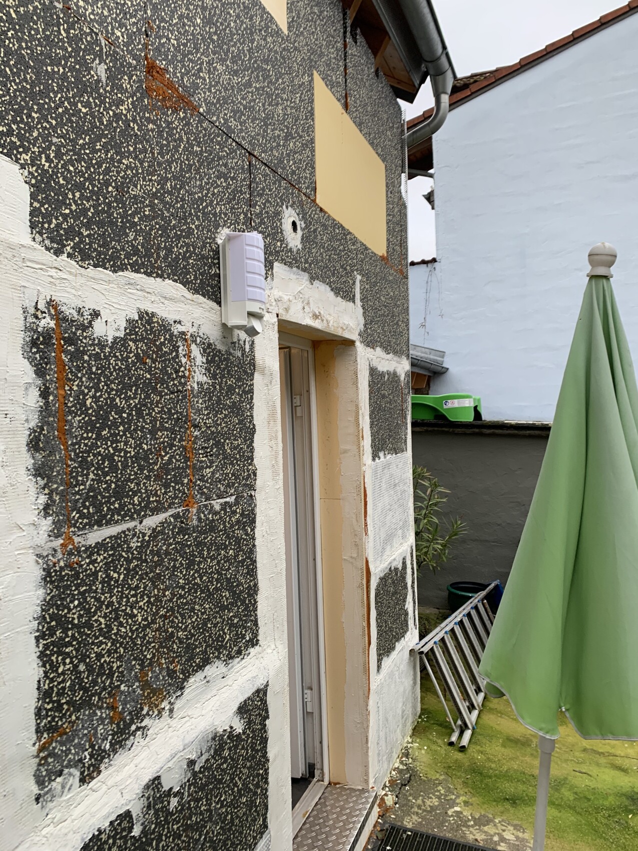 Außenwand mit sichtbarer Dämmung aus dunklen Platten, Türöffnung und Gartenutensilien