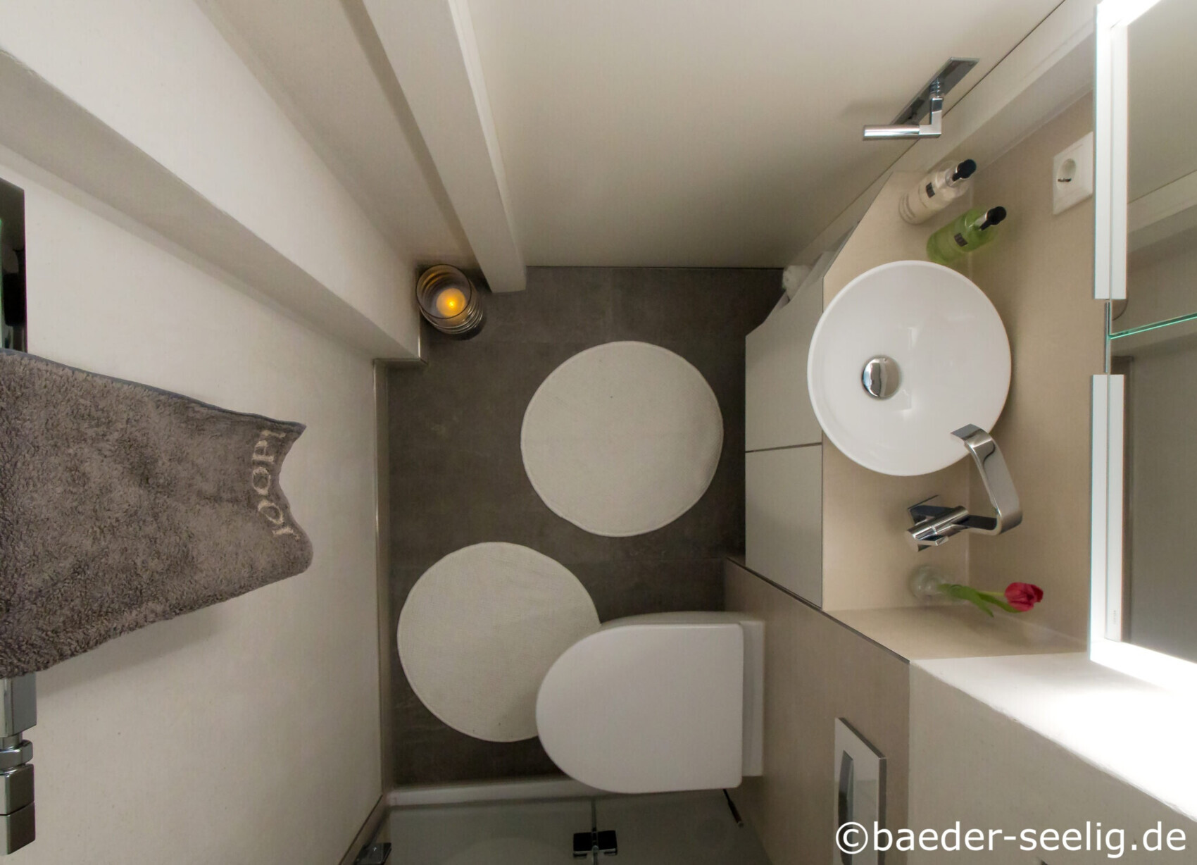Small bathroom from above: round washbasin, toilet, two round rugs, towel on the wall.