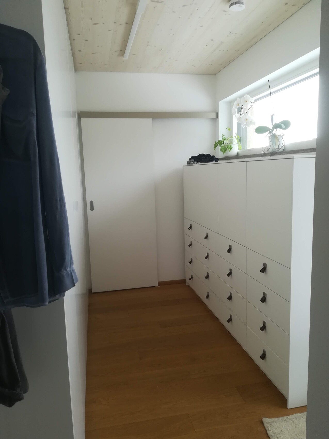Narrow wardrobe room with white cabinets on the right, window with plants, wooden floor.