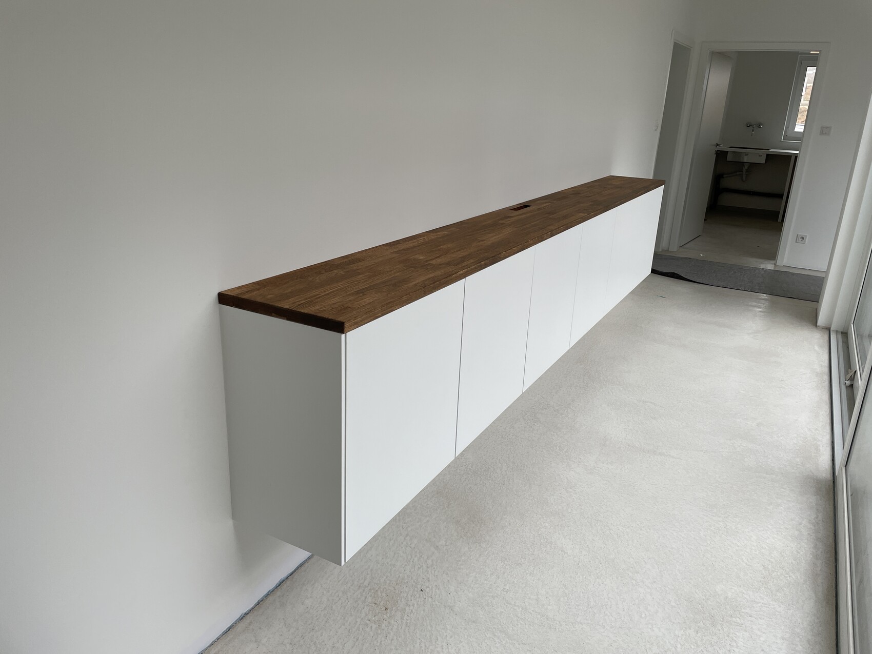 Long white cabinets with wood surface along the wall of a hallway; open bathroom in the background.