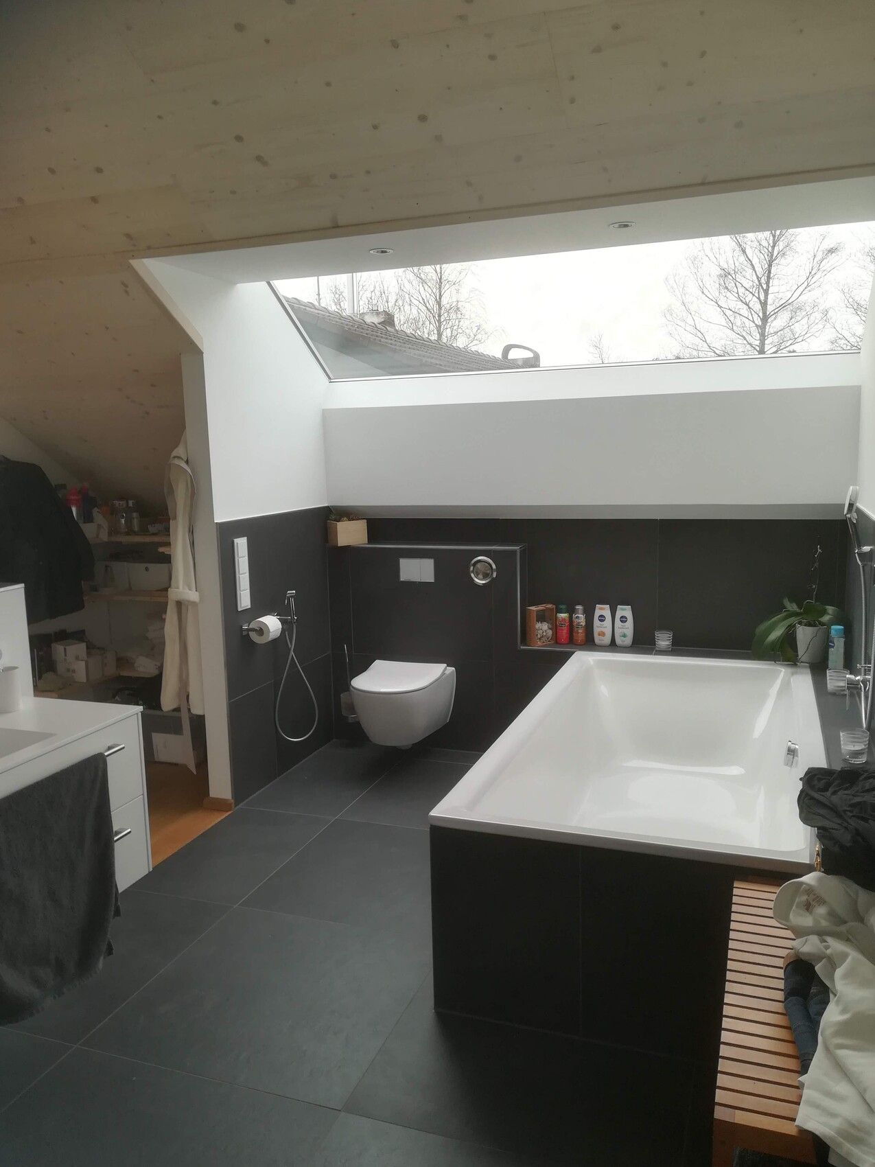 Modern bathroom in the attic with built-in bathtub, toilet, dark tiles, and skylight.