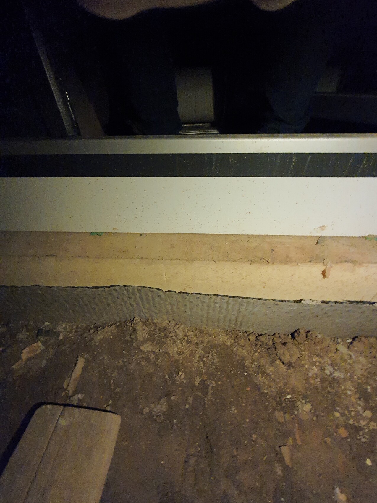 Unter einem dunklen Möbelstück: staubiger Boden, Erde und Holzreste, weiße Sockelleiste.