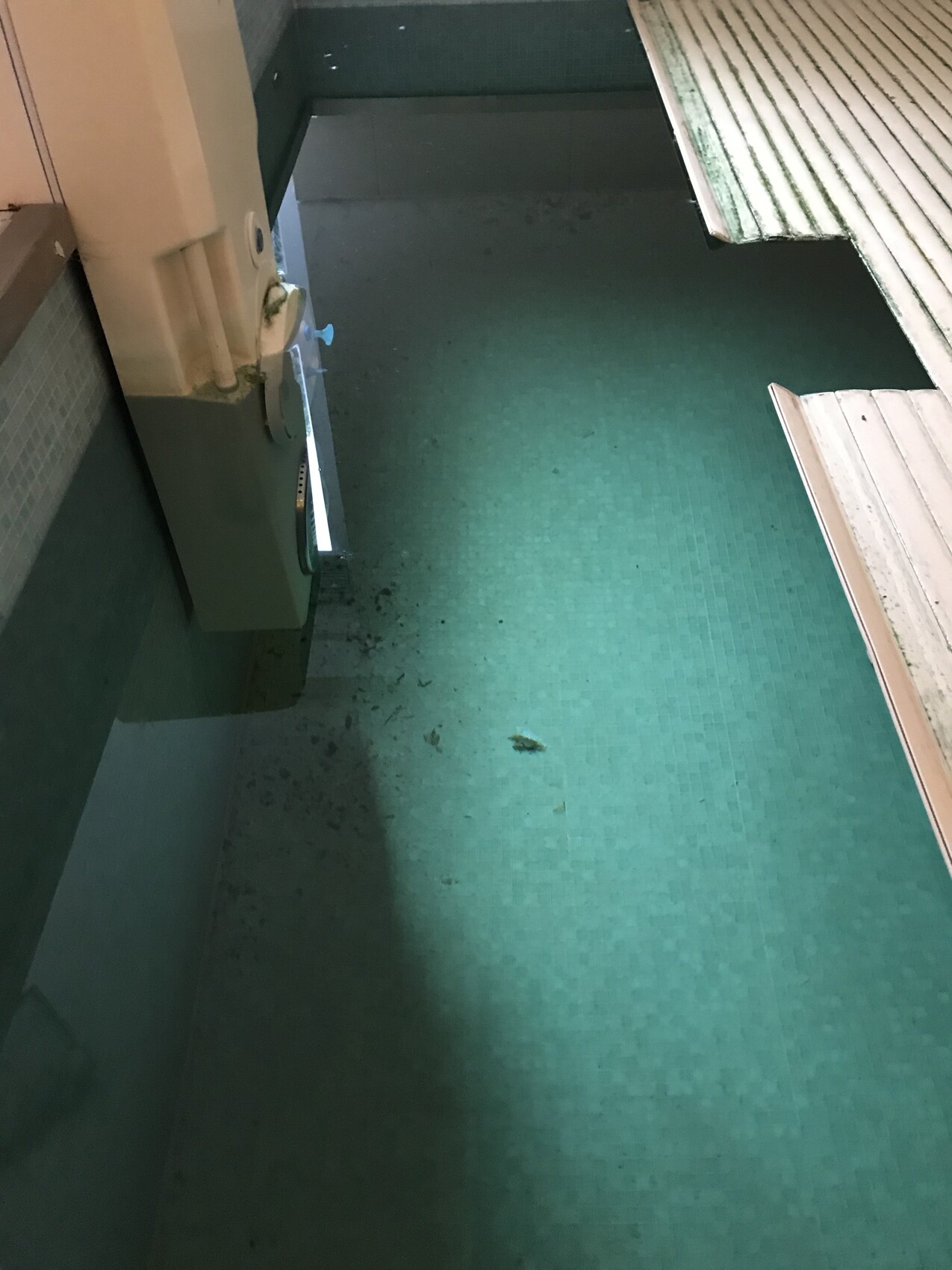 Green mosaic floor indoors; left wall with device, right side with wooden planks.
