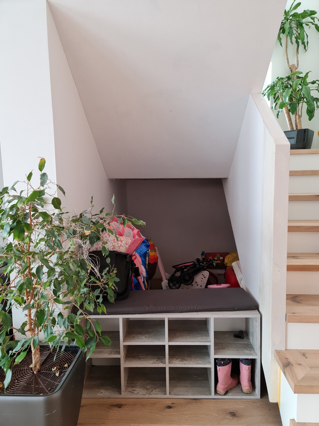 Bench under the staircase with compartments, shoes, toys; on the left a large plant.