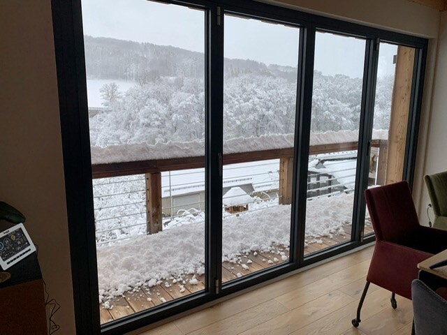 Snow-covered balcony behind large glass windows, view of snowy forest landscape.