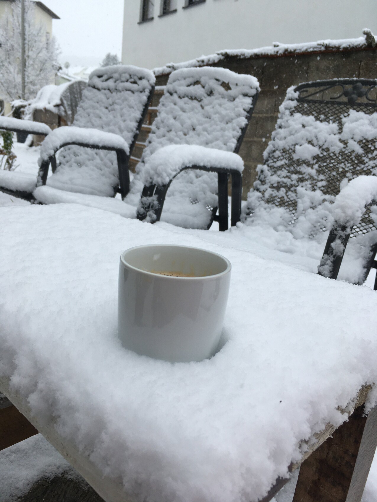 White coffee cup sitting on snow on an outdoor table; snow-covered garden chairs in the background.