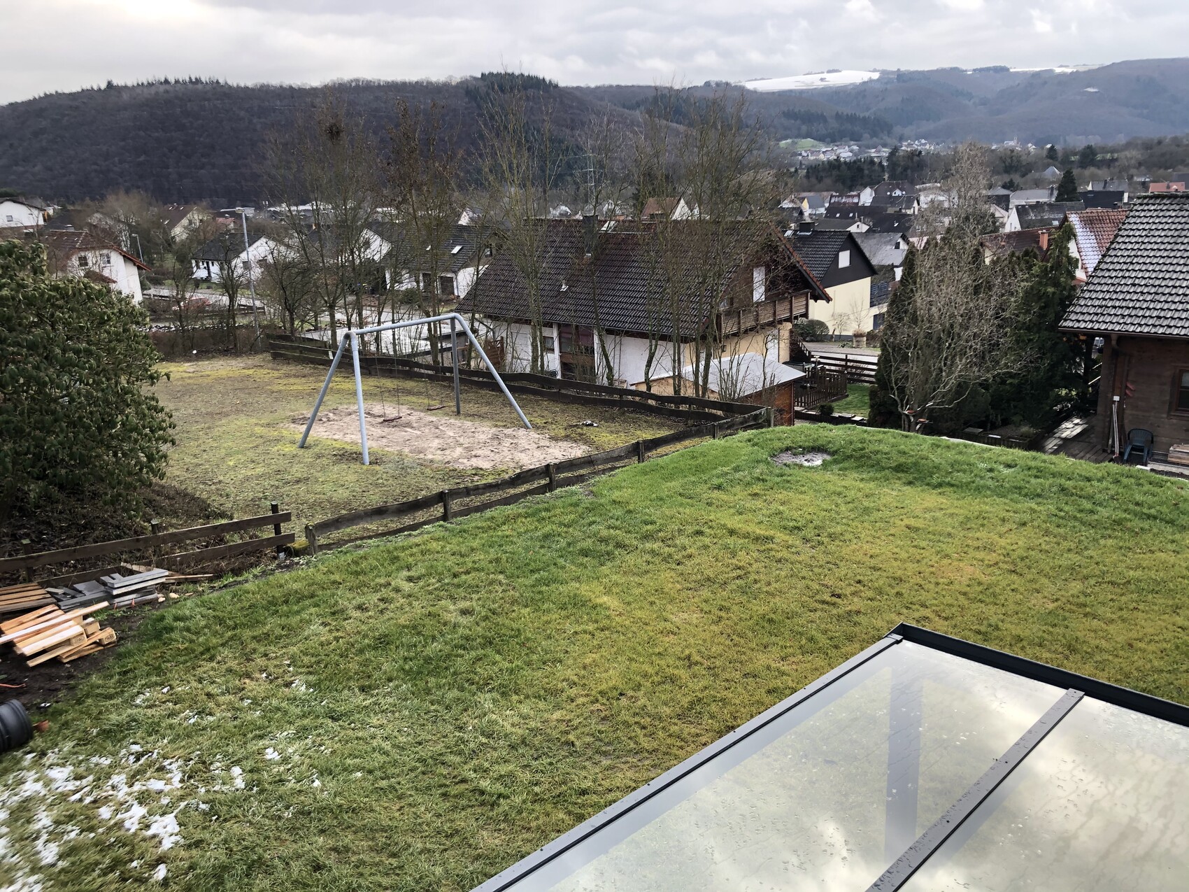 Garten mit Schaukel auf Sand, Zäune, Häuserreihen und hügelige Berglandschaft im Hintergrund.