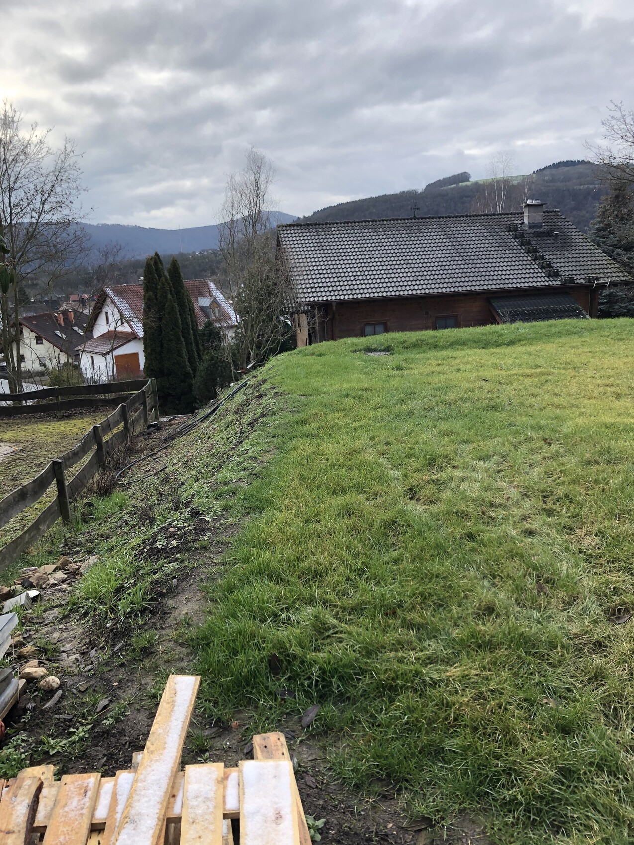 Grüner Hang mit Bauholz am Boden, Holzzaun links, Haus mit Ziegeldach und Hügeln im Hintergrund.