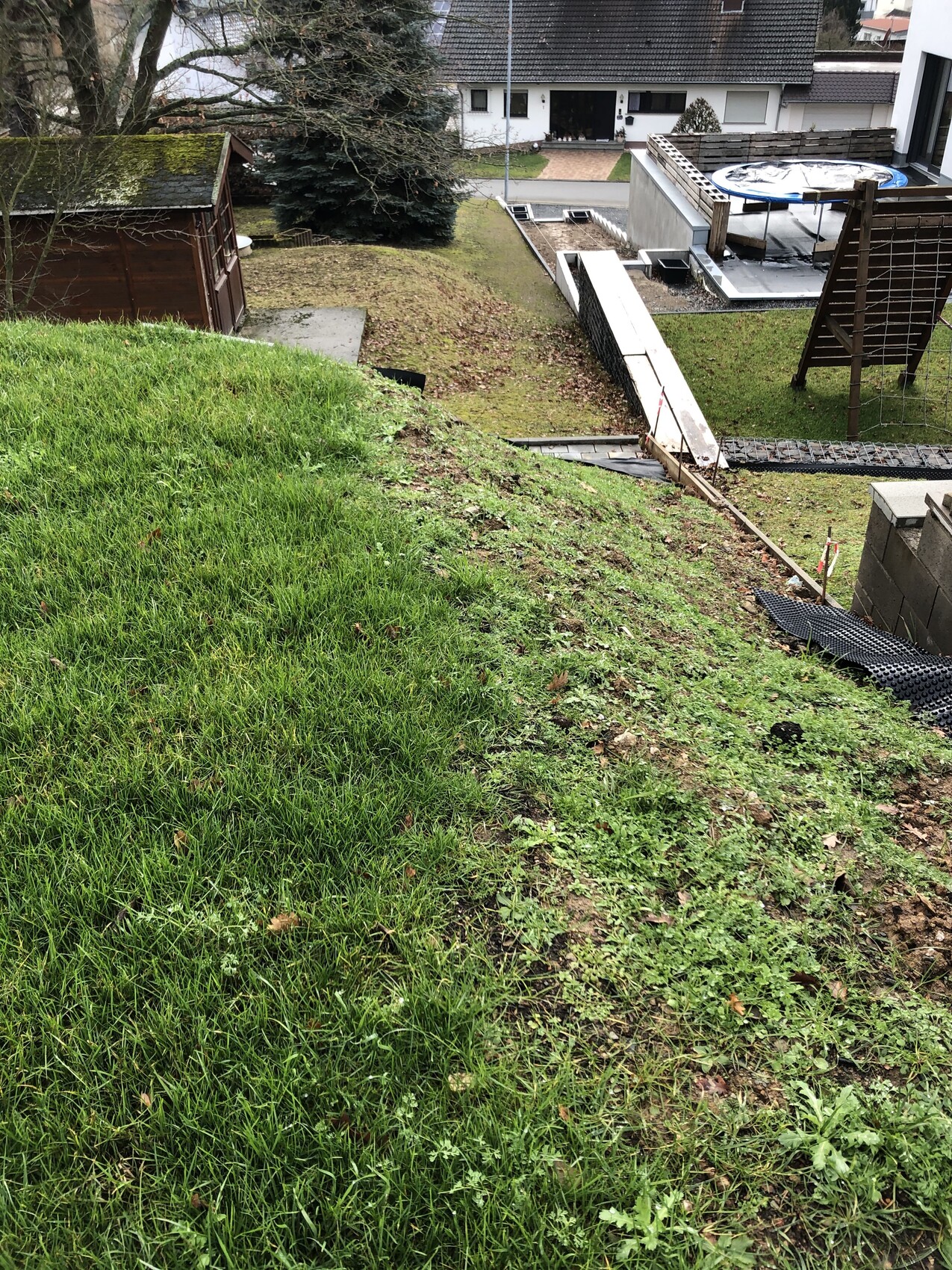 Steiler Gartenhang mit grünem Gras, linkes Holzhaus, rechts Trampolin, Rampe und Häuser im Hintergrund