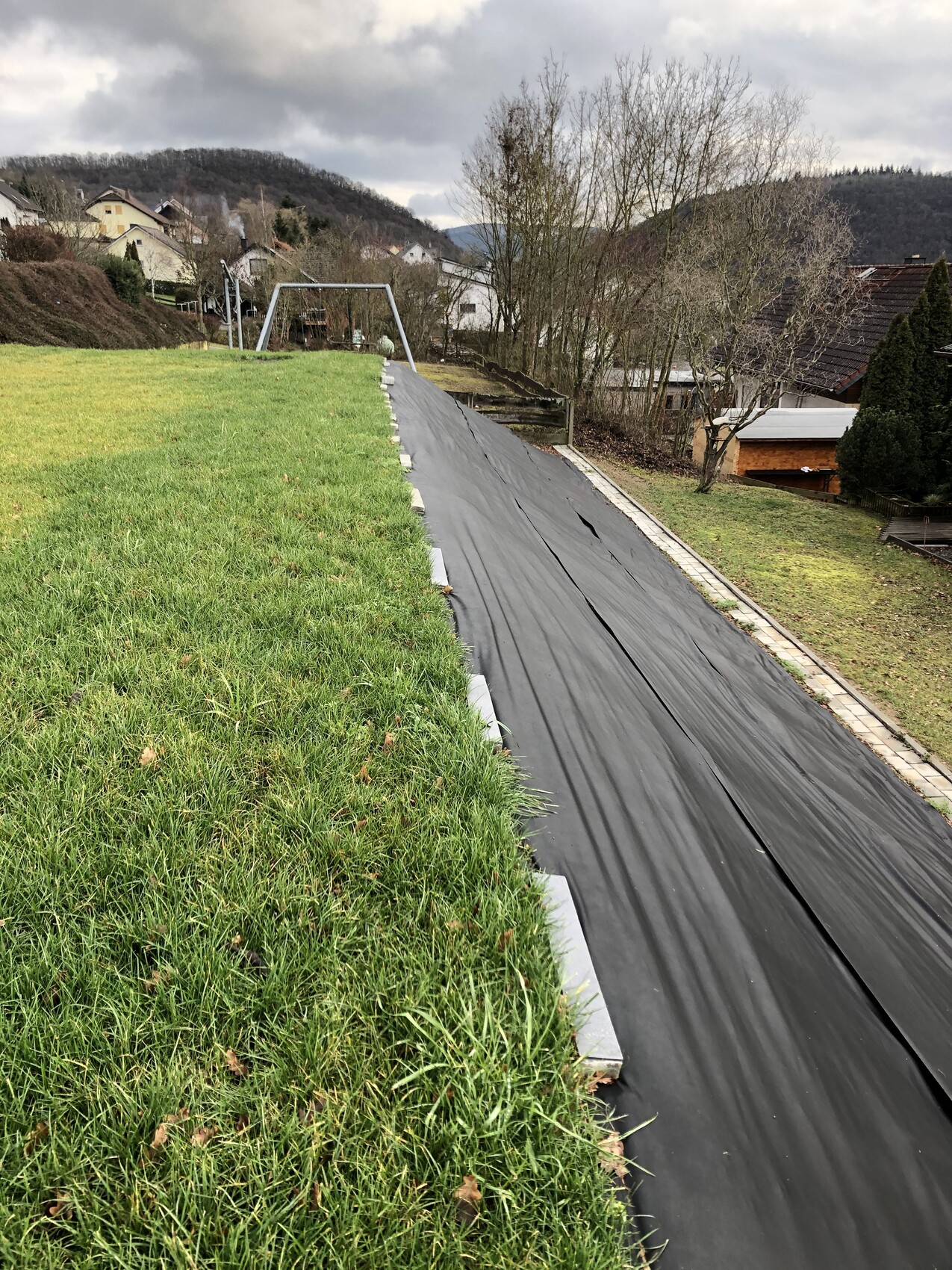 Grüne Böschung links, mit schwarzer Folie bedeckt; rechts gepflasterter Weg, Dorf im Hintergrund