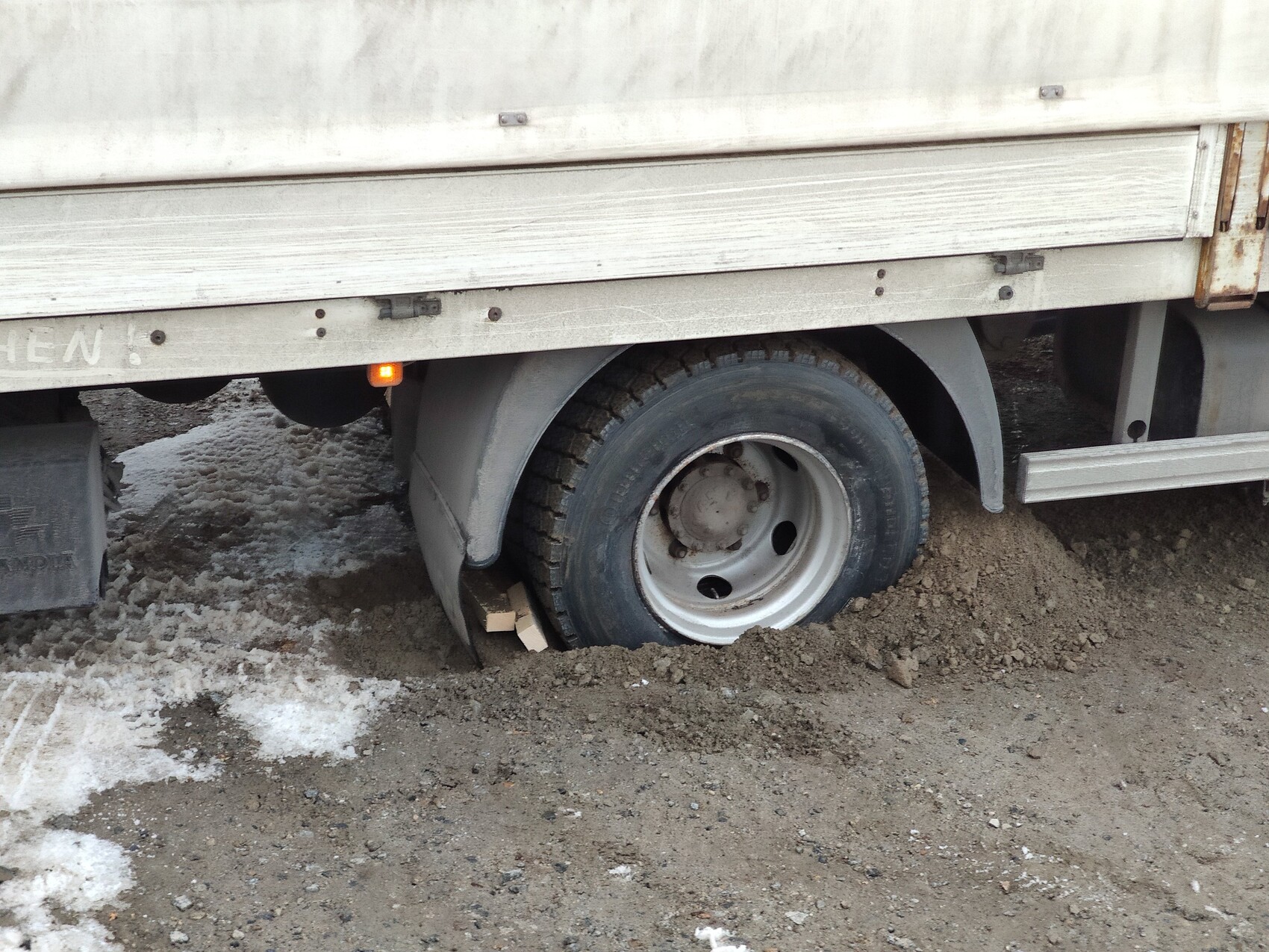 Truck trailer with wheel stuck in mud, wooden block underneath, dusty construction site environment.