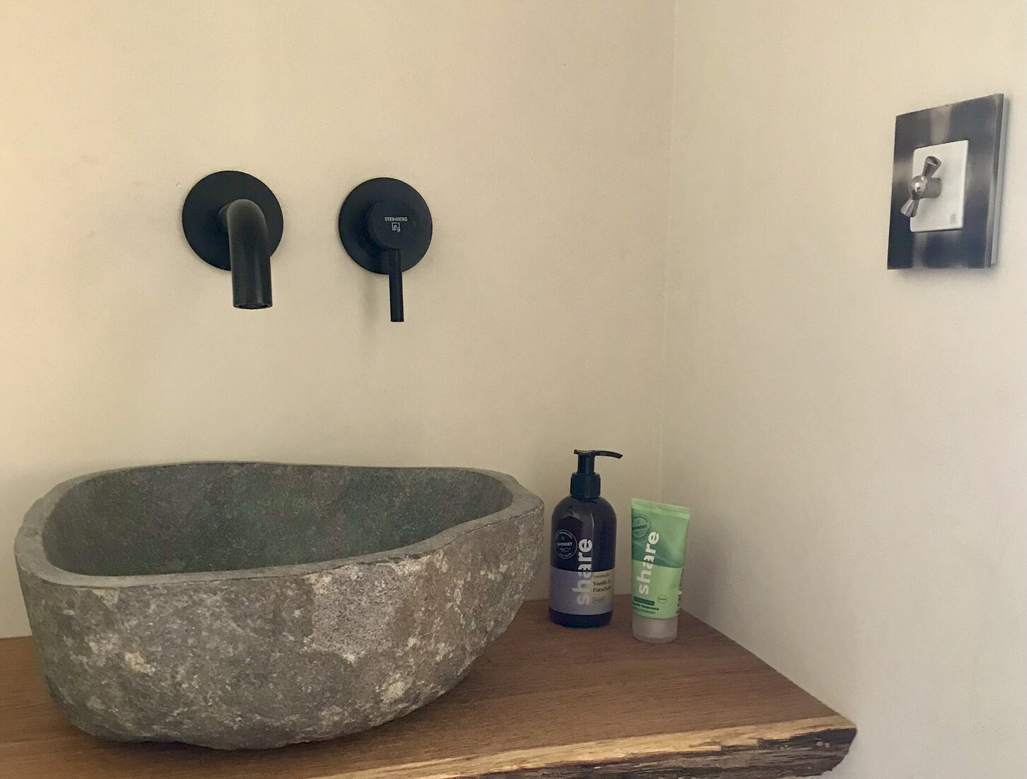 Stone washbasin on wooden table with black wall-mounted faucet and soap dispenser.