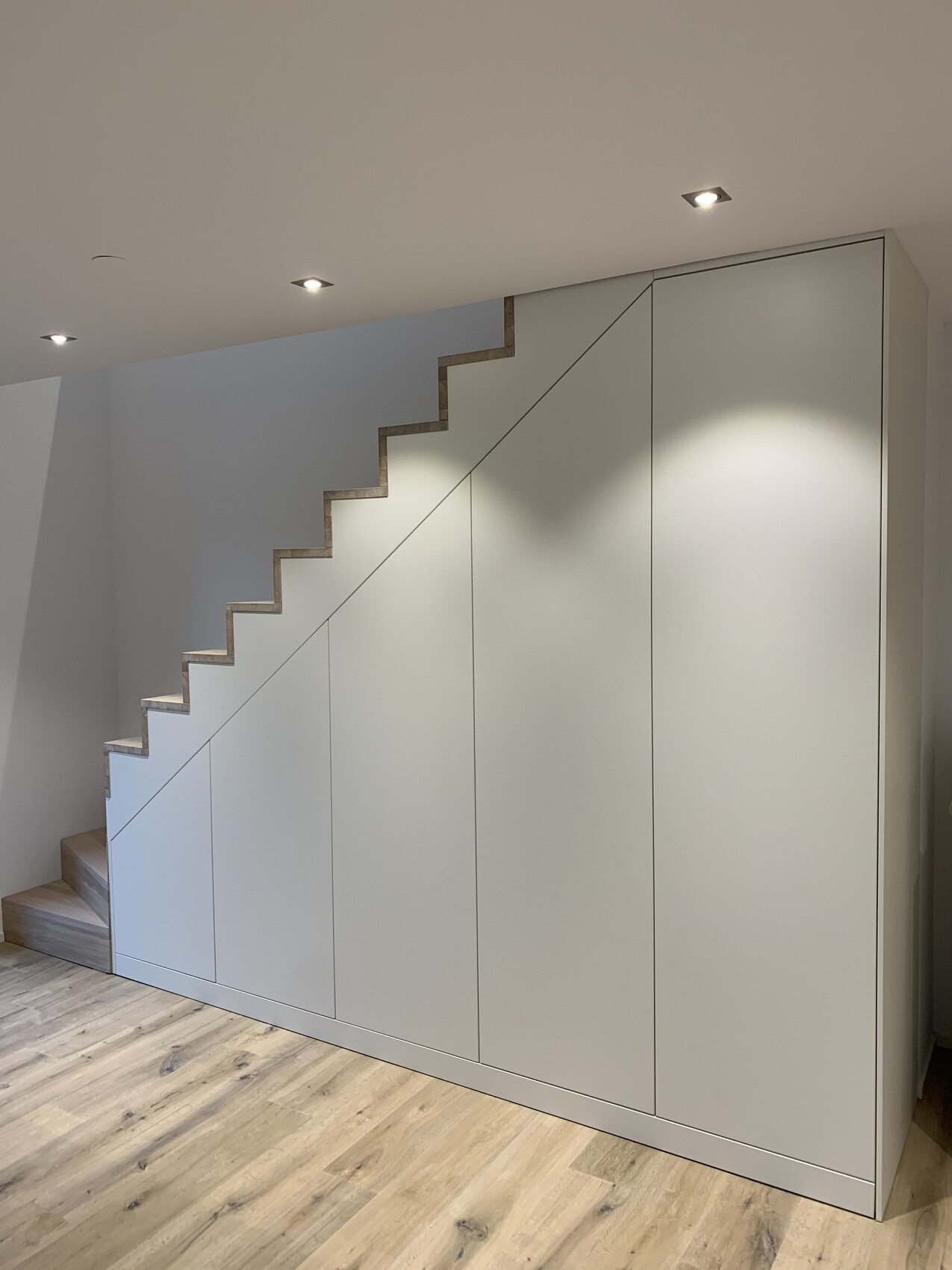 Modern interior view: white built-in cabinets with diagonally running wooden staircase, hardwood floor.