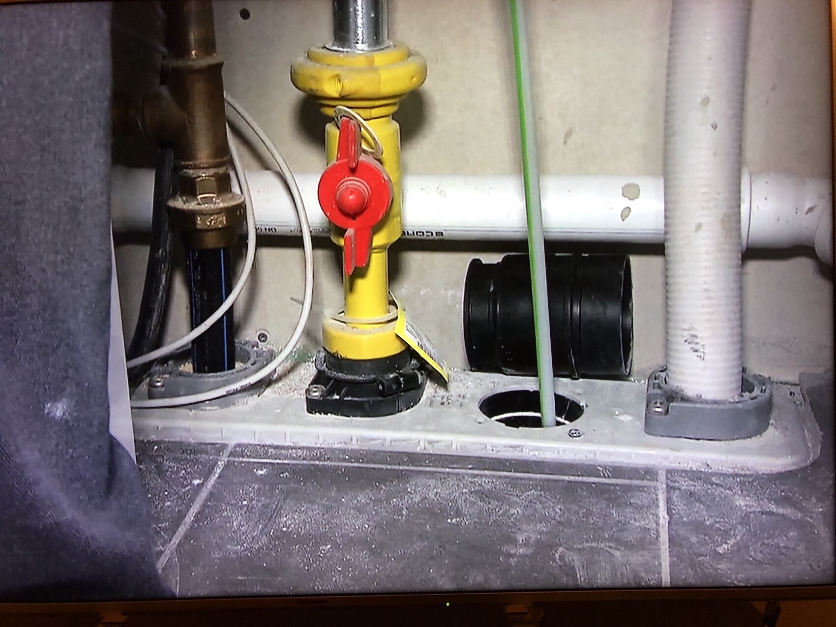 Yellow shut-off valve with red handle in front of pipes, cables, and floor opening.
