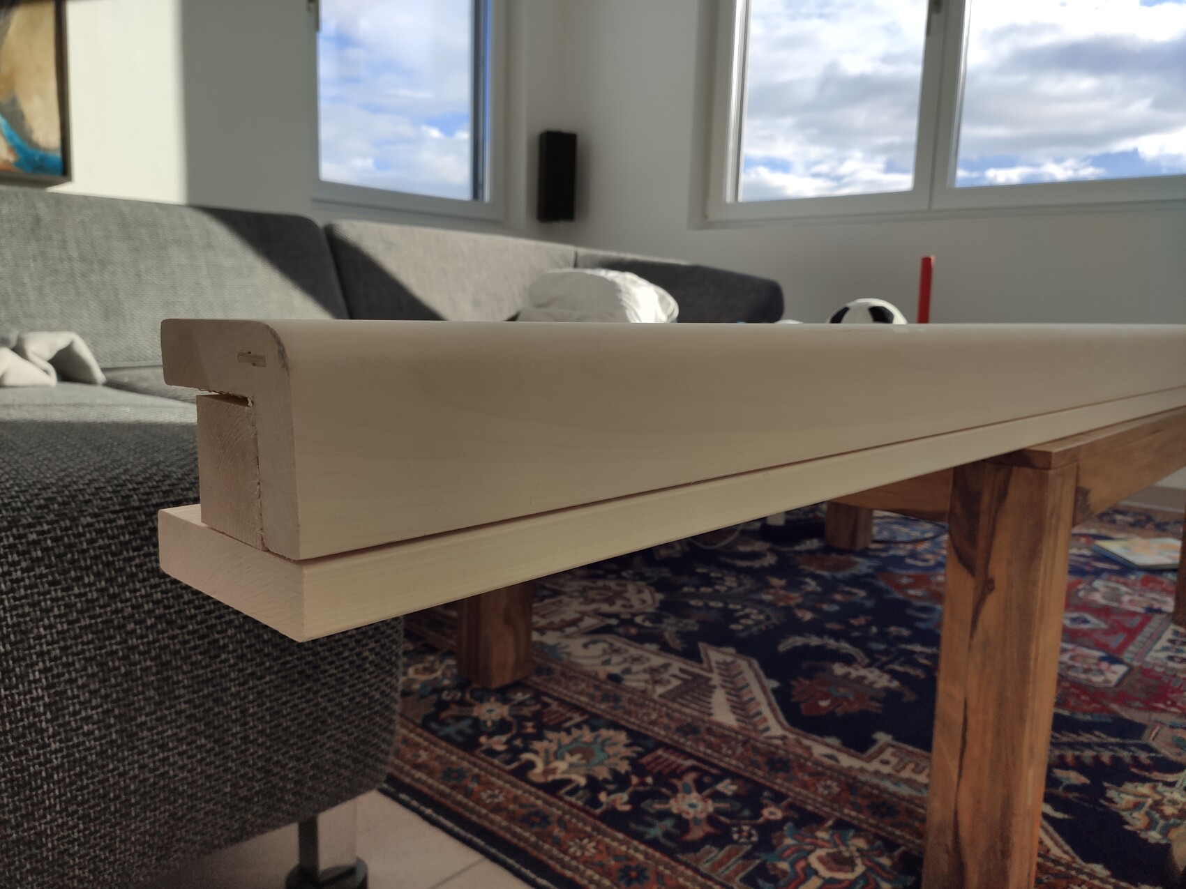 Elongated light wooden board lying across two wooden beams; sofa and window in the background.