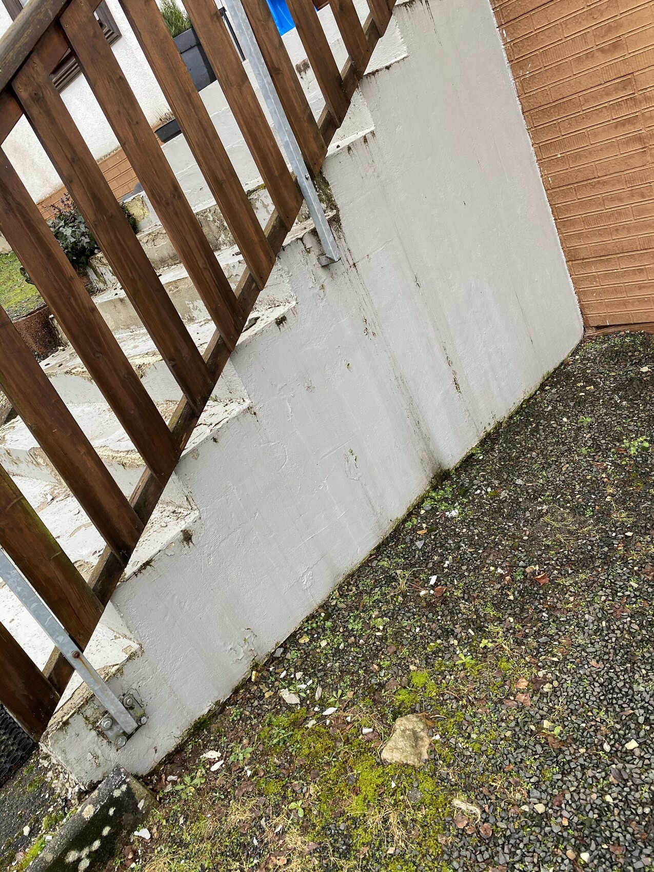 Outdoor steps with brown wooden fence next to a white wall, beside brick wall and mossy gravel ground.