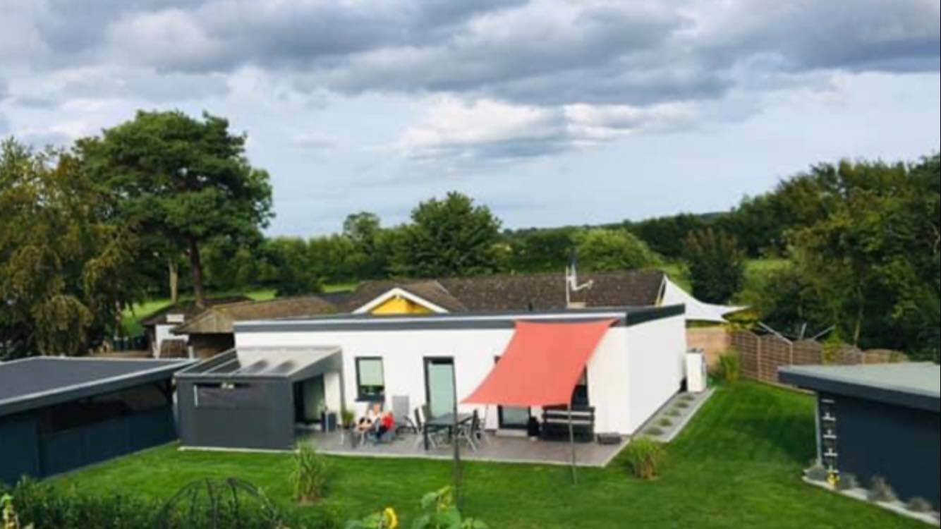 Moderne weiße Einfamilienhaus mit rotem Sonnensegel, Terrasse und Sitzgruppe im Garten.