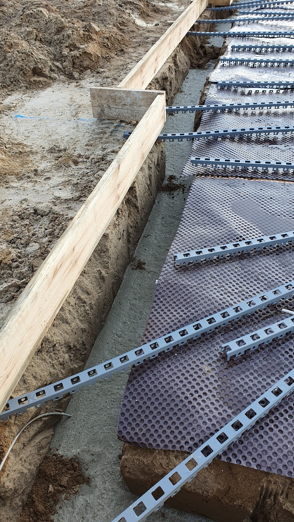 Excavation pit with wooden formwork, perforated metal channels lying across, and anti-slip mat.