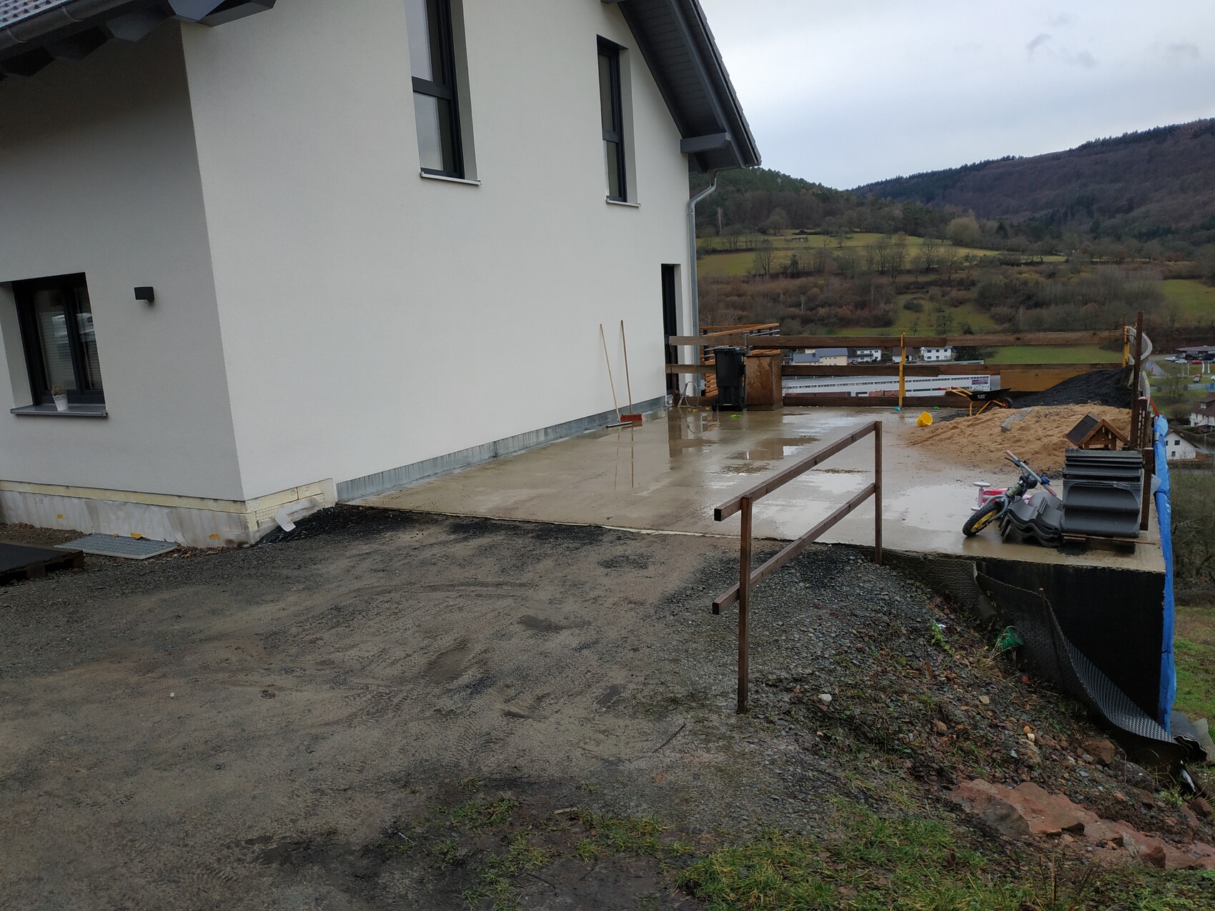 Außenansicht eines Neubaus; Terrasse, regennasse Fläche, Bauutensilien, Geländer, Berglandschaft.