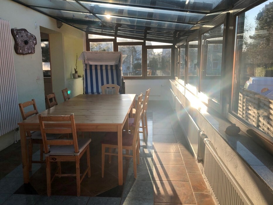Sunlit veranda with large wooden table, chairs, and glass roof, view outside.