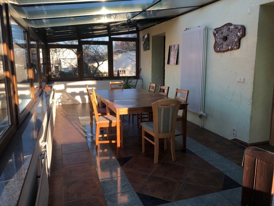 Bright glass conservatory with large wooden table and chairs, sunlight coming through windows from the left.