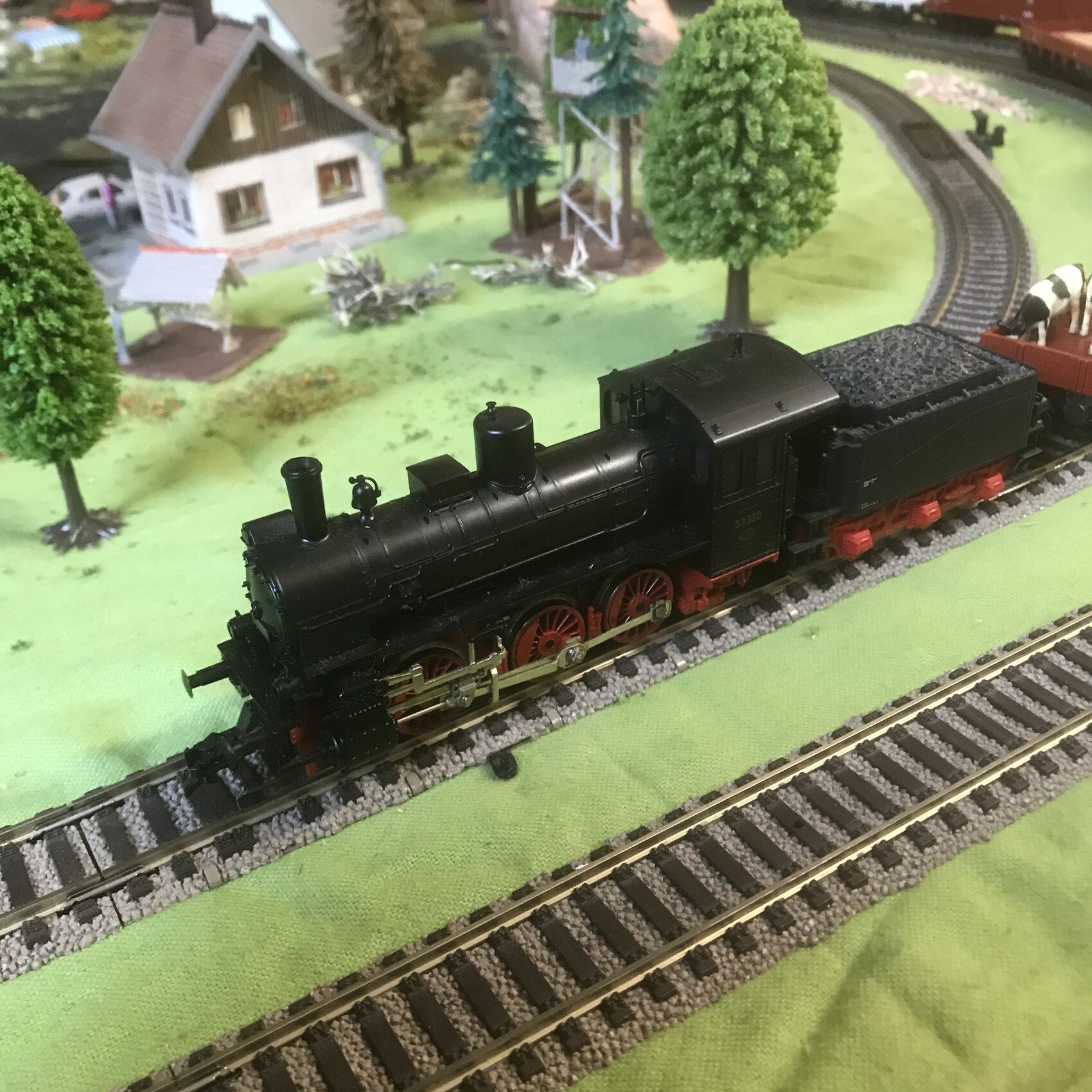 Black model steam locomotive with red chassis pulling coal cars on model railway through landscape.