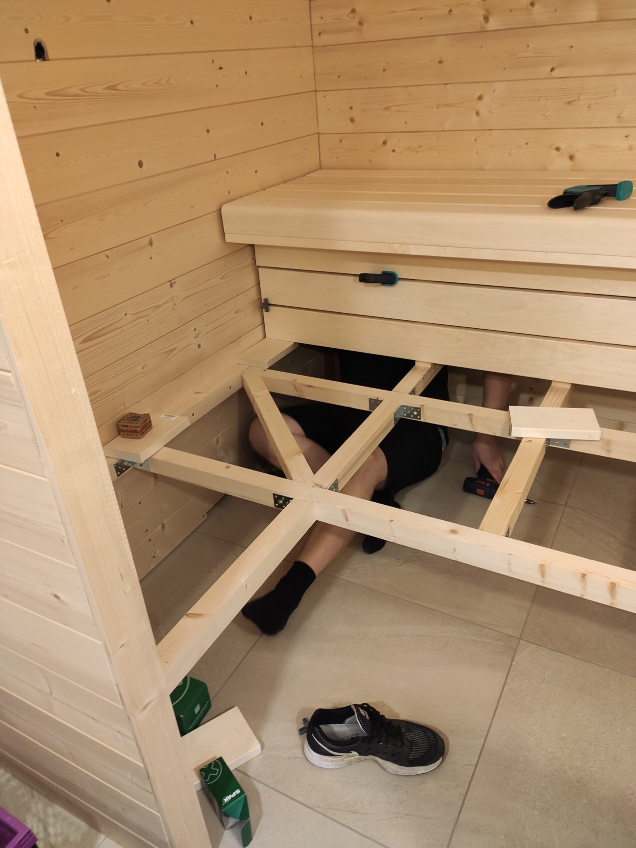 Person working under a wooden frame structure in a wooden room; tools lying nearby.