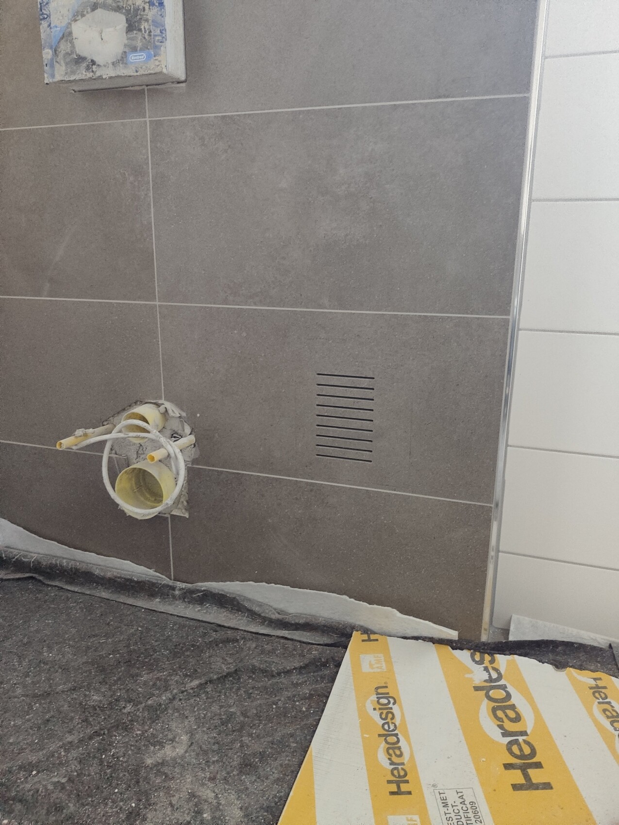 Construction site wall with gray tiles, exposed yellow sanitary pipe, and ventilation slot.