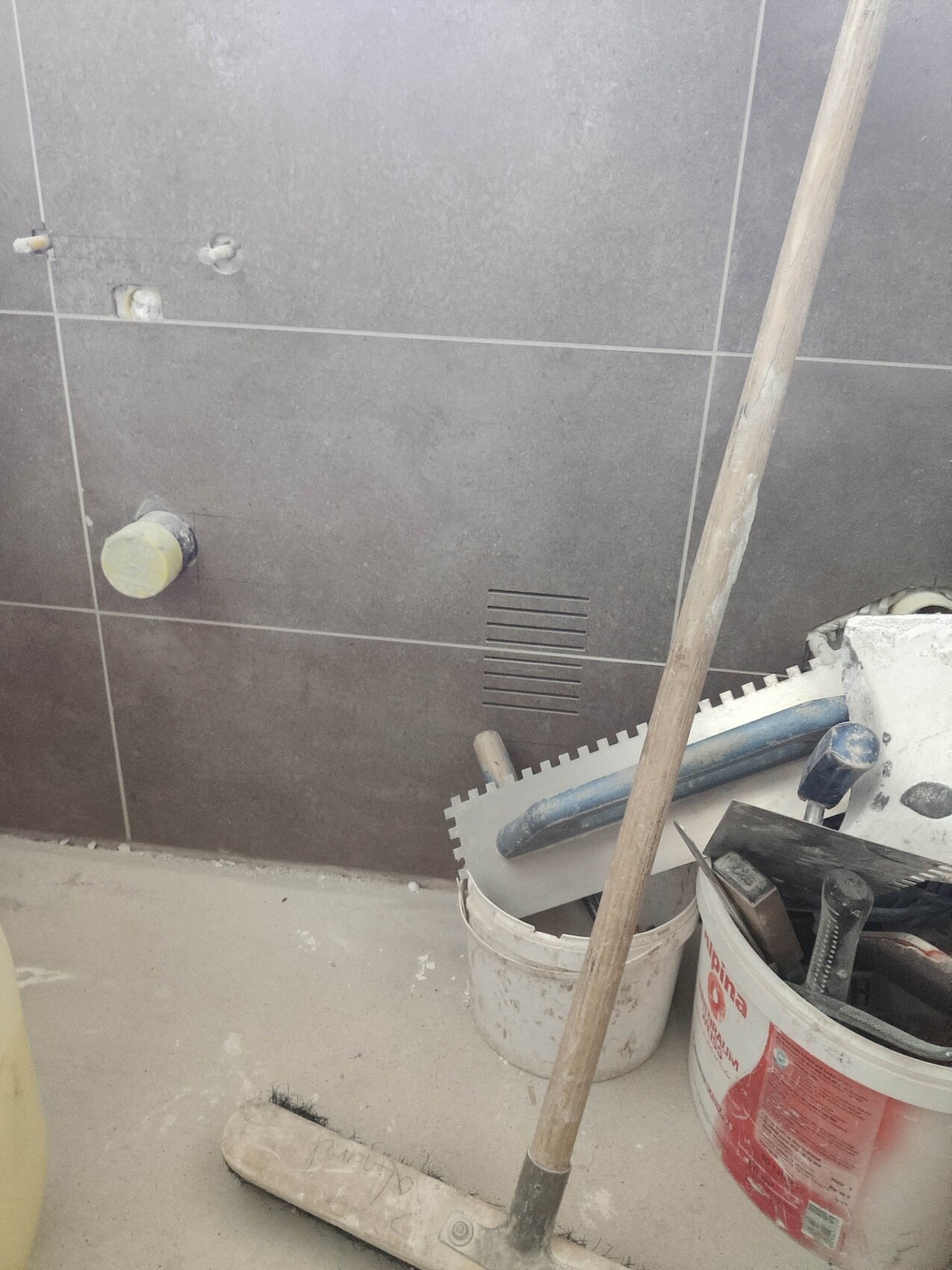 Gray tiled room under construction: bucket with tools, broom handle, and tile tool.