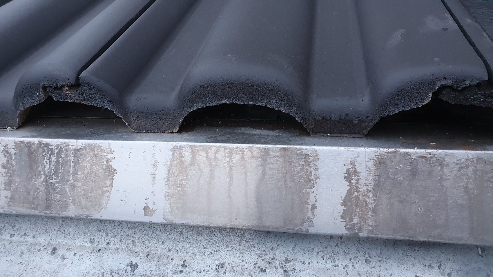 Close-up of dark roof tiles with a broken edge; underneath, a dirty metal trim.