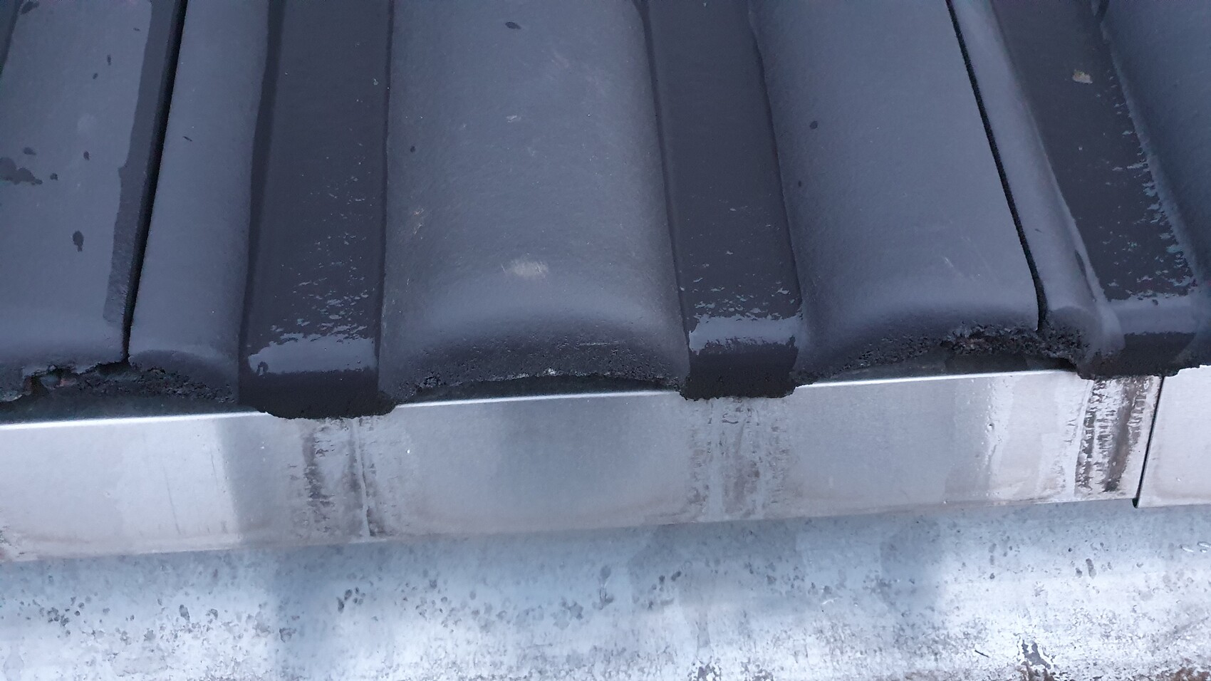 Close-up of dark blue roof tile with moisture, below a metal edge.