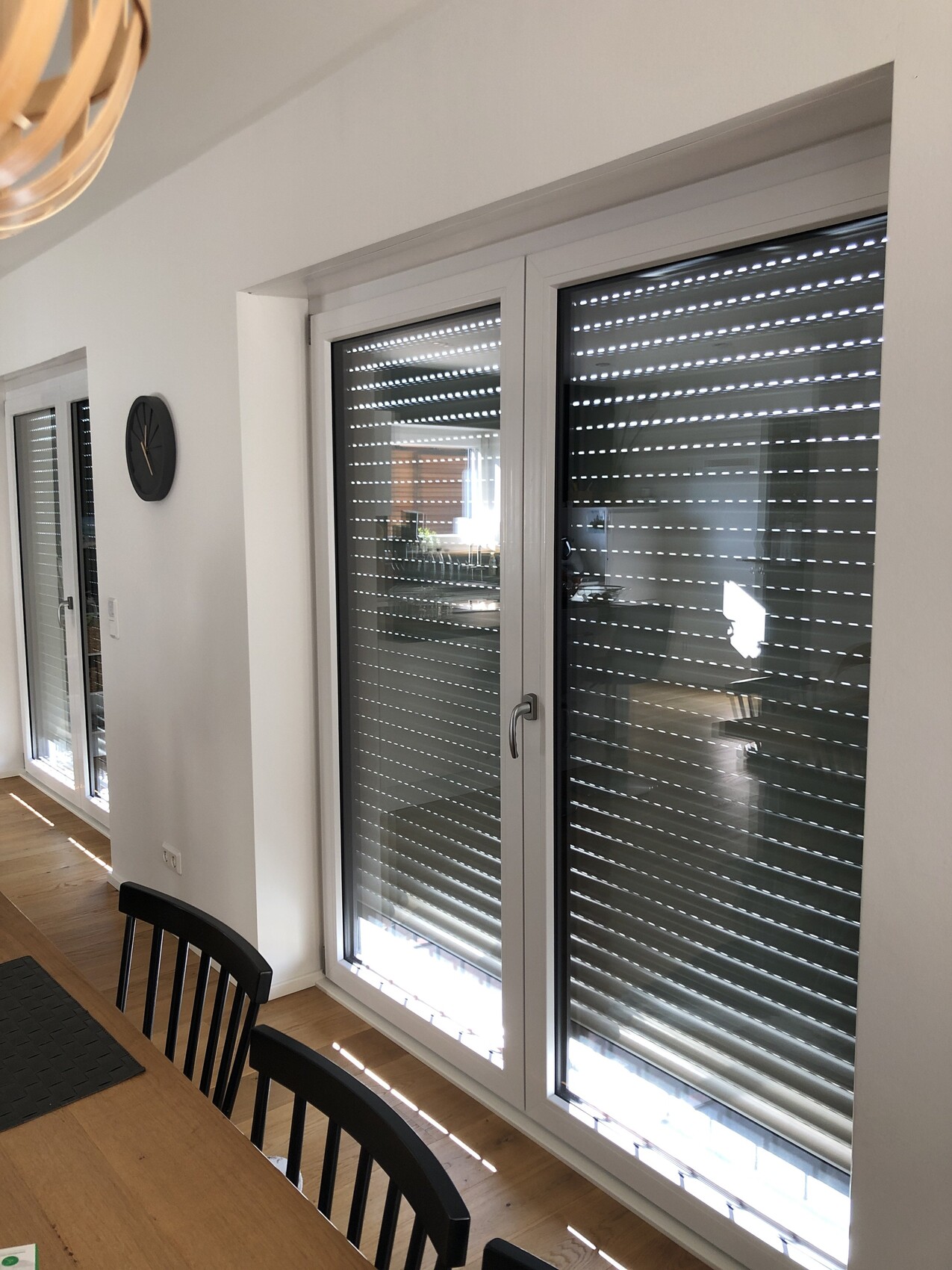 Interior view: dining area with large windows and roller shutters, wooden floor, table and chairs.
