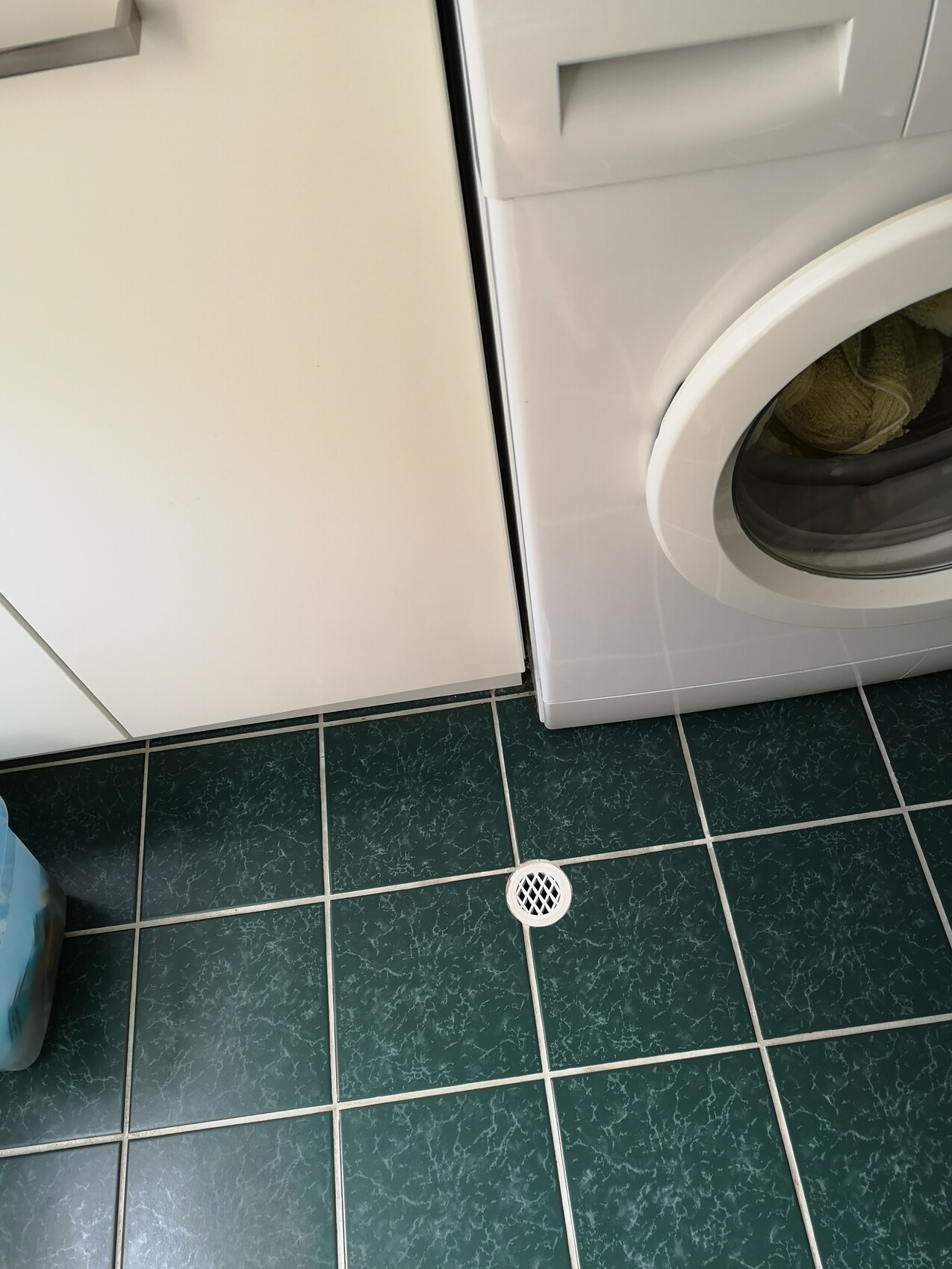 Front-loading washing machine next to a white cabinet on a green tiled floor with a floor drain.