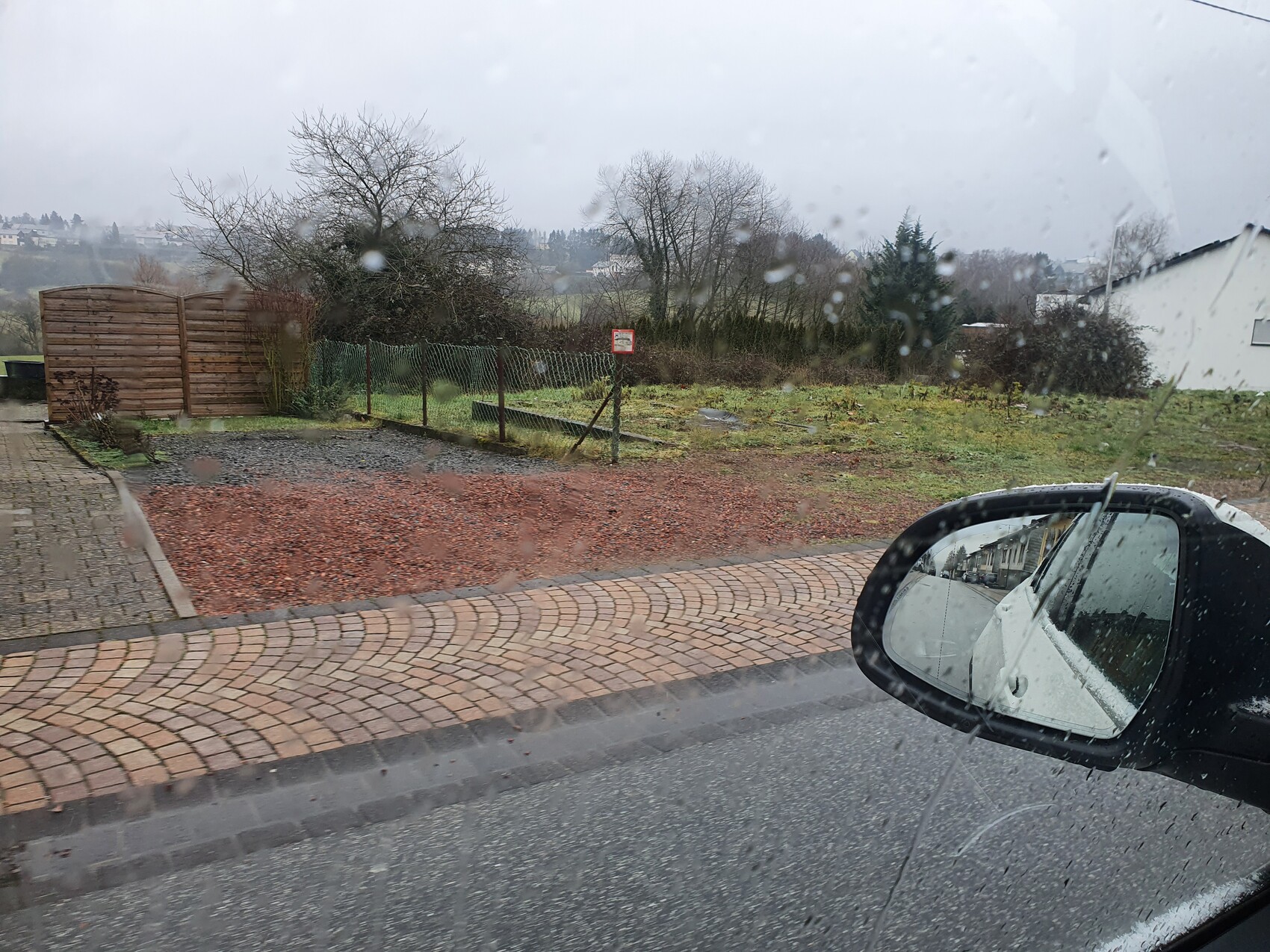 View from car in rain: wet windshield, cobblestone, garden fence, mirror shows street scene.