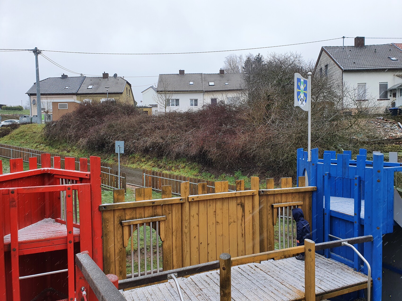 Colorful wooden playground: red, brown, and blue structures; houses in the background; child in hoodie.