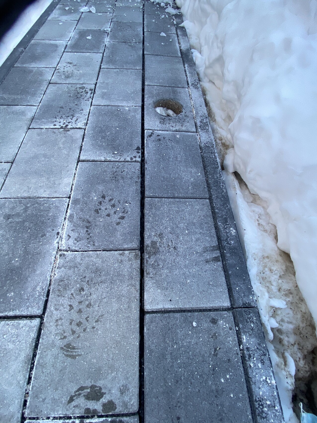 Paved path made of square stone slabs, snow on the right, footprints and hole in the paving.