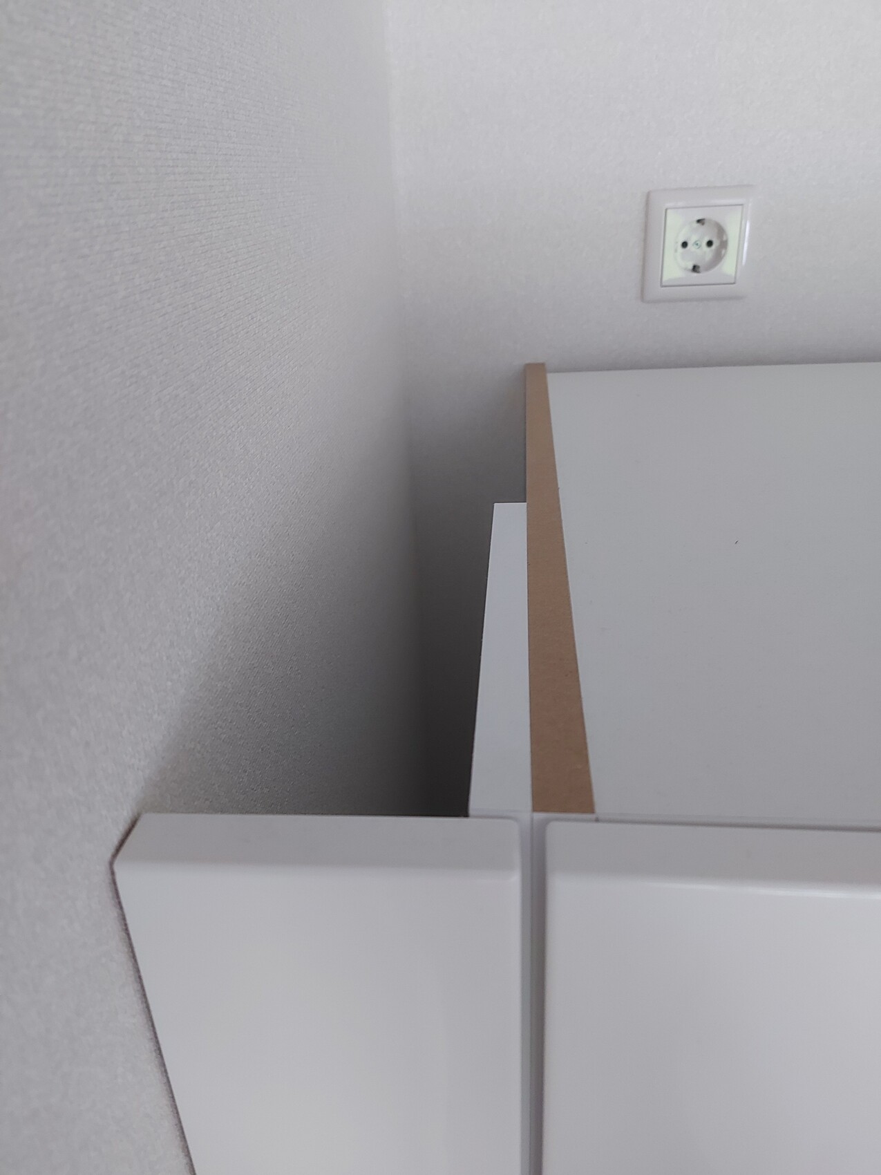 Corner view of a white cabinet with a brown edge in front of a gray wall; power outlet visible.